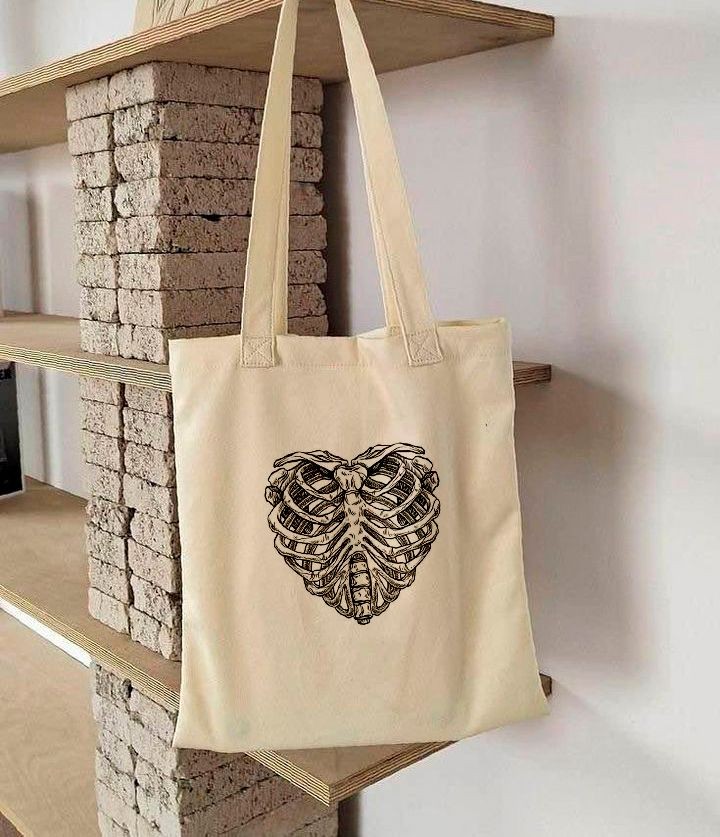 Beige tote bag with heart-shaped ribcage design hanging on a wooden shelf.