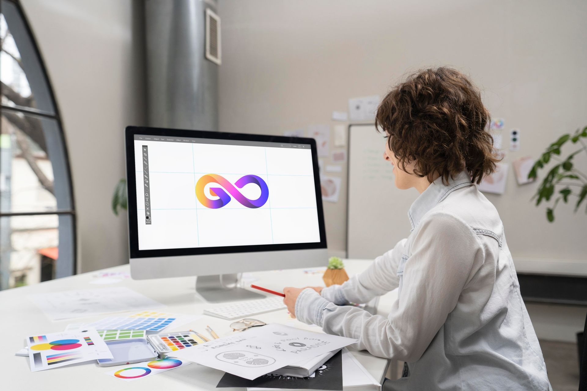 Designer working on computer with infinity logo on screen, surrounded by sketches and color palettes.