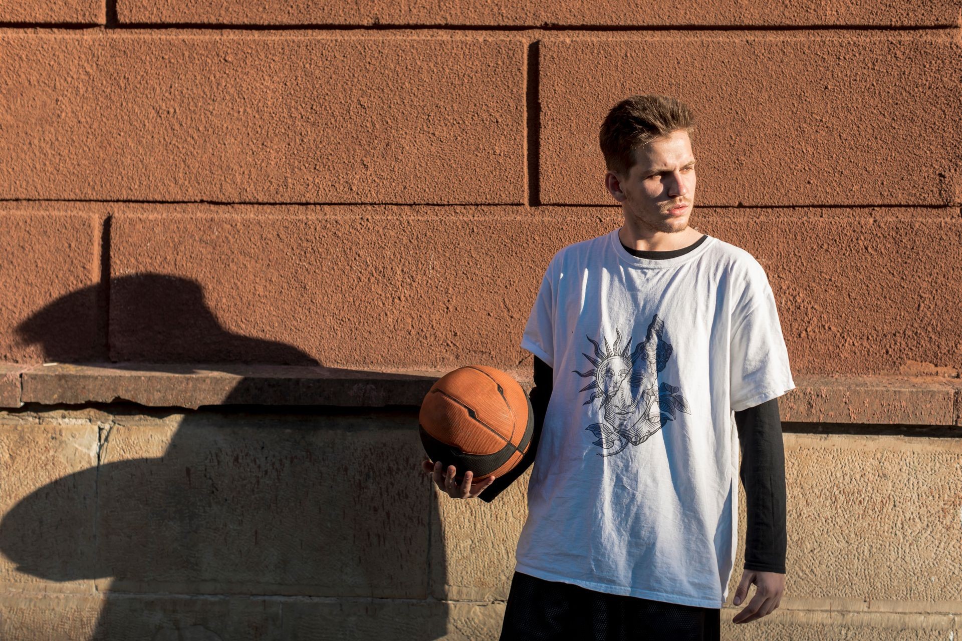 Person wearing a white graphic t-shirt holding a basketball in front of a textured brick wall.